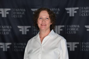 A woman with curly hair smiles in front of a backdrop featuring the 'Farm & Forge Club' logo. She wears a white shirt.